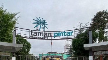 Gerbang masuk Taman Pintar Yogyakarta berbentuk lengkung dengan tulisan taman pintar, dengan bilik pengunjung kosong di sisi kiri dan kanan, pagar di bagian depan, langit mendung, dan poster protokol kesehatan kecil di bilik kanan.