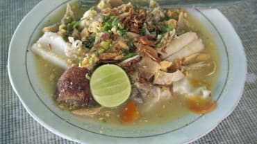 Semangkuk Soto Banjar berisi kuah bening kekuningan, suwiran ayam, potongan lontong, perkedel kentang, bawang goreng, seledri, dan irisan jeruk nipis.