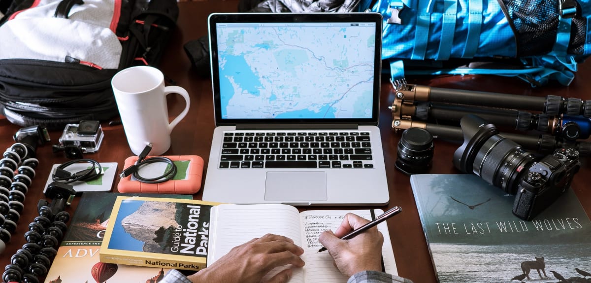 Meja dengan laptop menampilkan peta, buku perjalanan, kamera, dan tangan yang menulis catatan