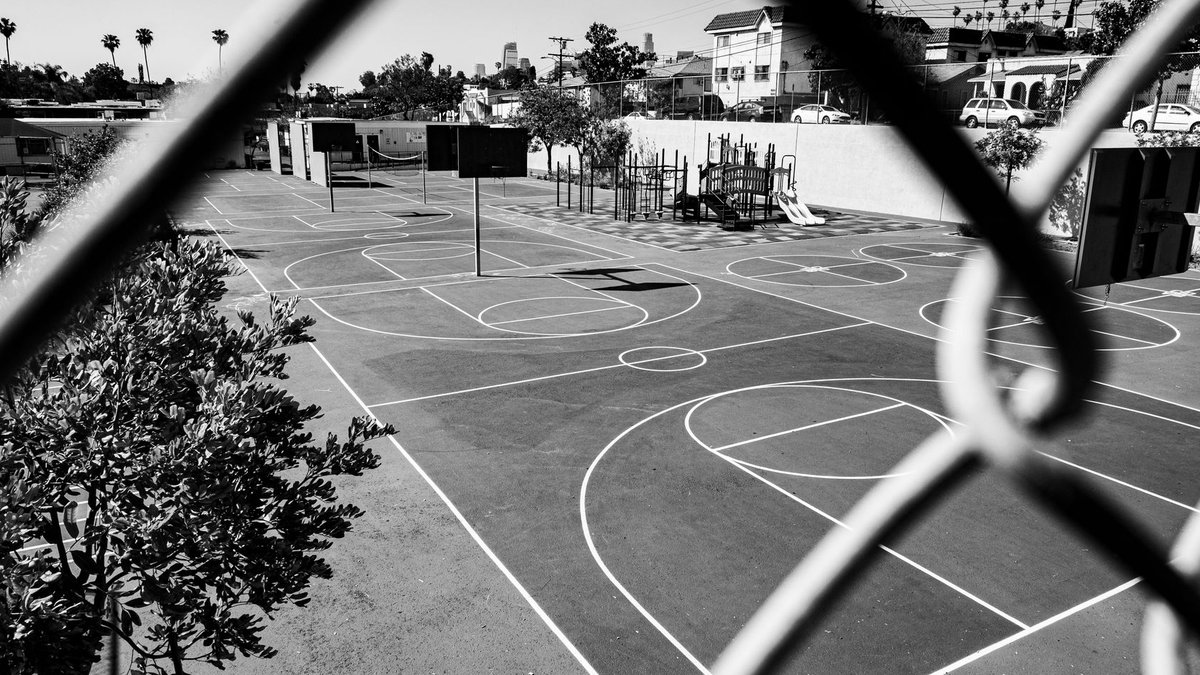 Black and white image of an outdoor basketball court viewed through a chain-link fence.