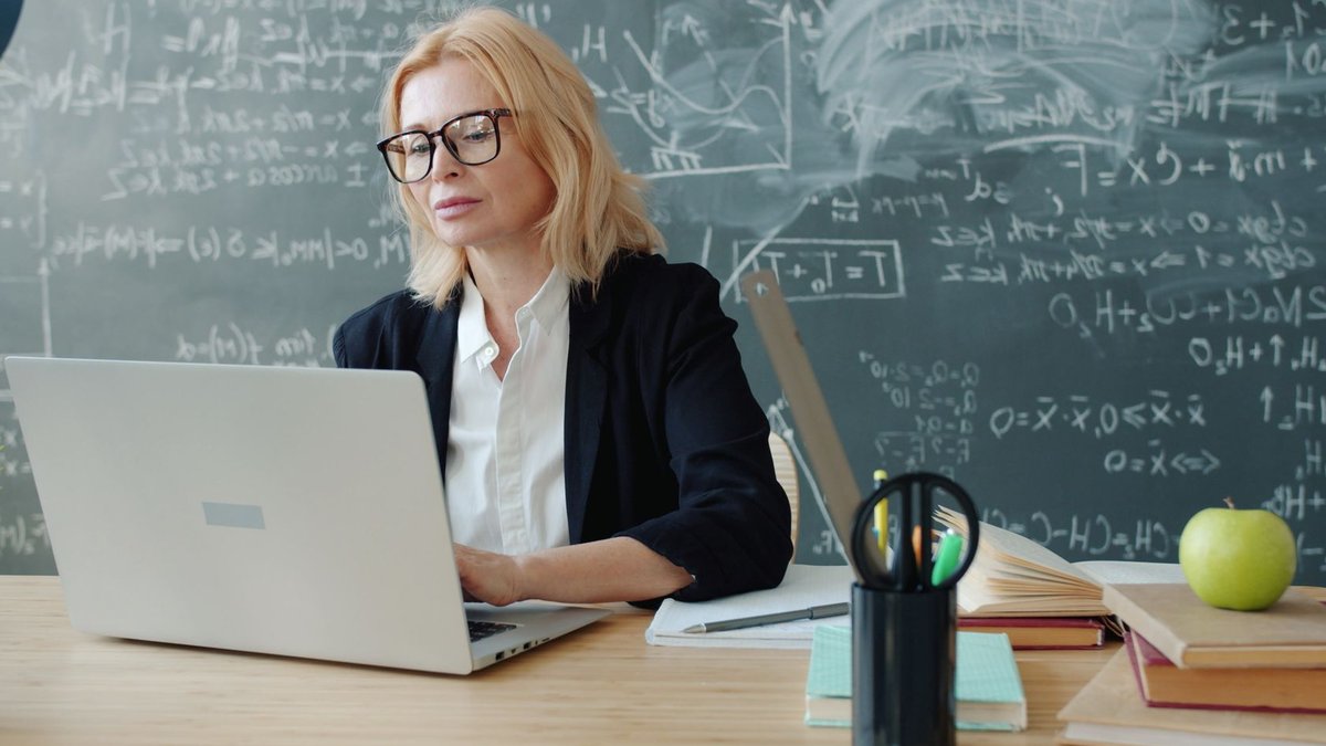 HR Leaders Weigh Study, Work Paths for Youth Teacher working on a laptop in a classroom with chalkboard equations, focused and professional.