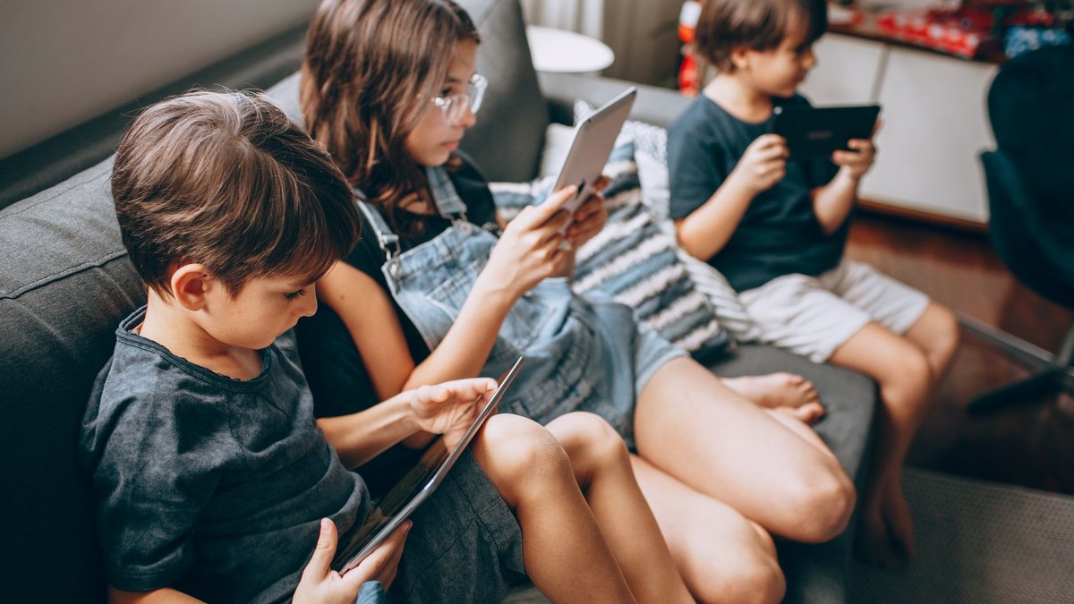 Three children using digital tablets while sitting together on a cozy sofa, enjoying technology indoors.