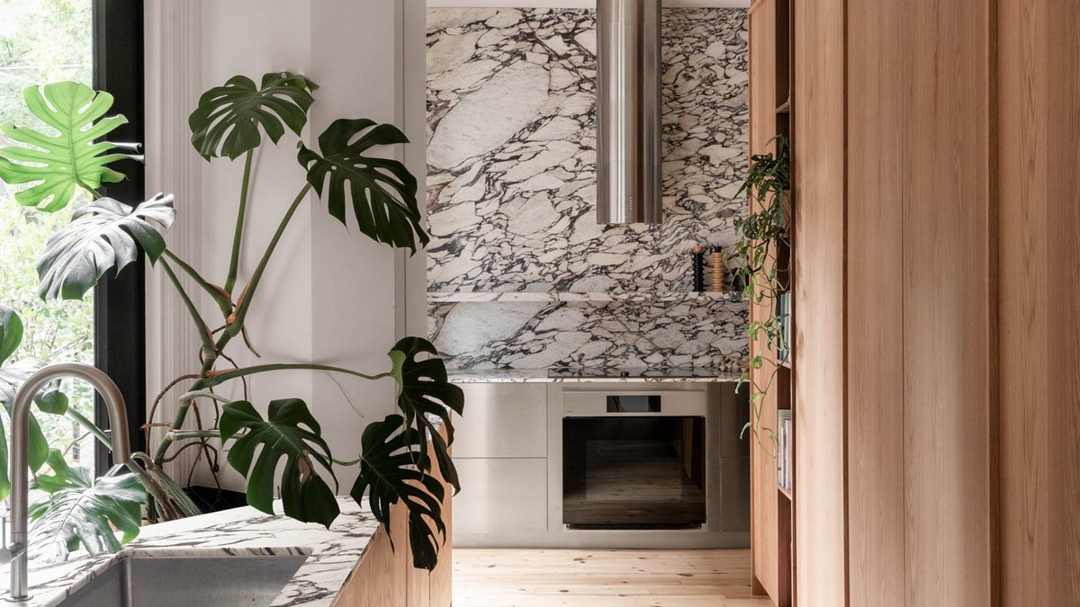Montreal Home Gets Modern Renovation Modern kitchen with marble backsplash and countertop, stainless steel oven, wood cabinetry, large windows, and a monstera pla