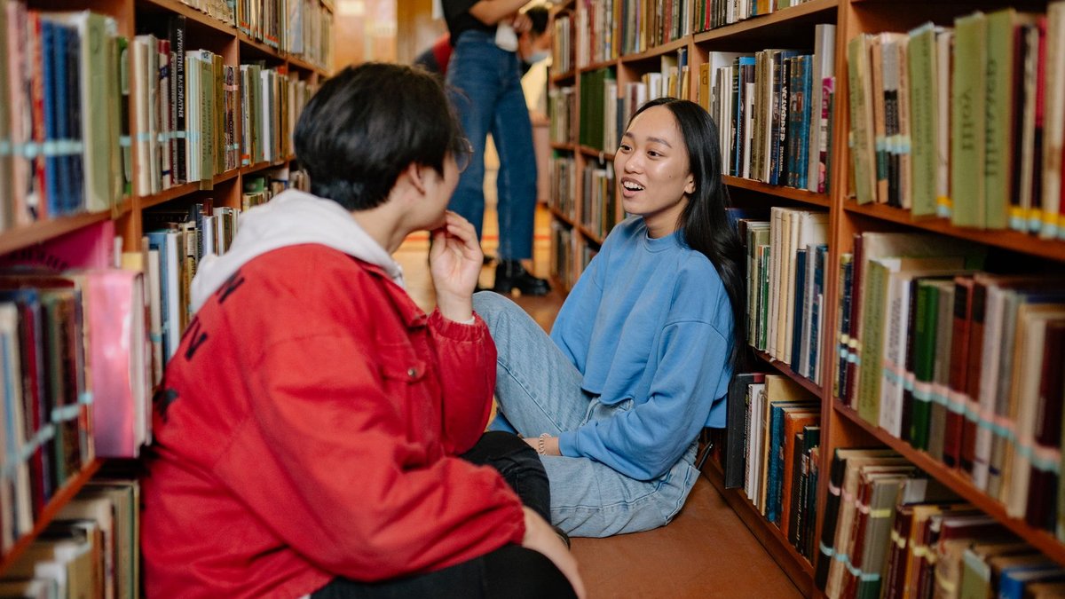 Two students having a conversation in a university library aisle.