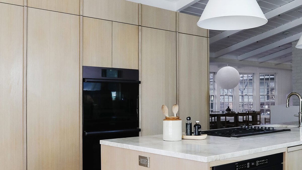 Designer hides kitchen appliances in seamless Lake Tahoe home Modern kitchen with light wood cabinets, black built-in oven, marble island with cooktop, and tableware on the counter; white