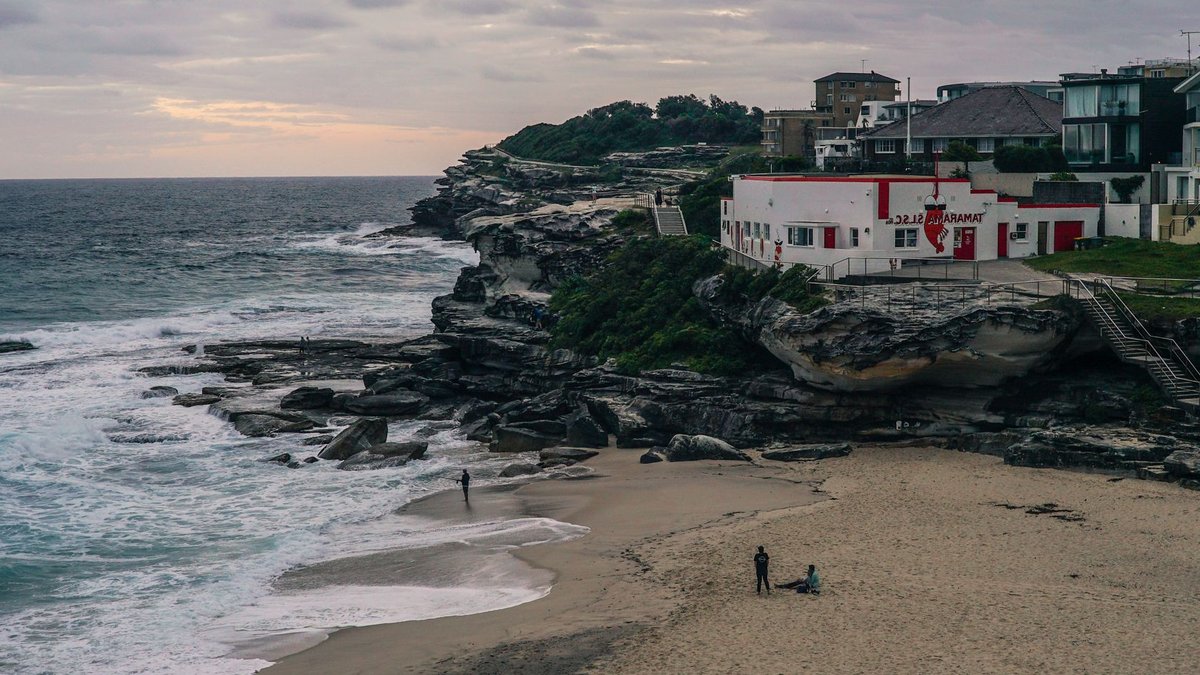 Bondi Backpackers to become Australia's first music hotel Tamarama Beach with cliffside houses under a cloudy sky, waves crashing on the shore.