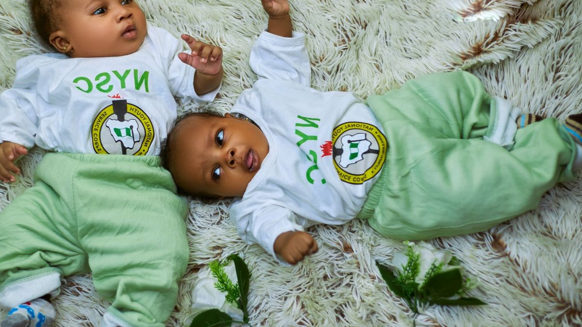 Adorable babies in NYSC attire lying on a cozy rug, representing innocence and national pride.