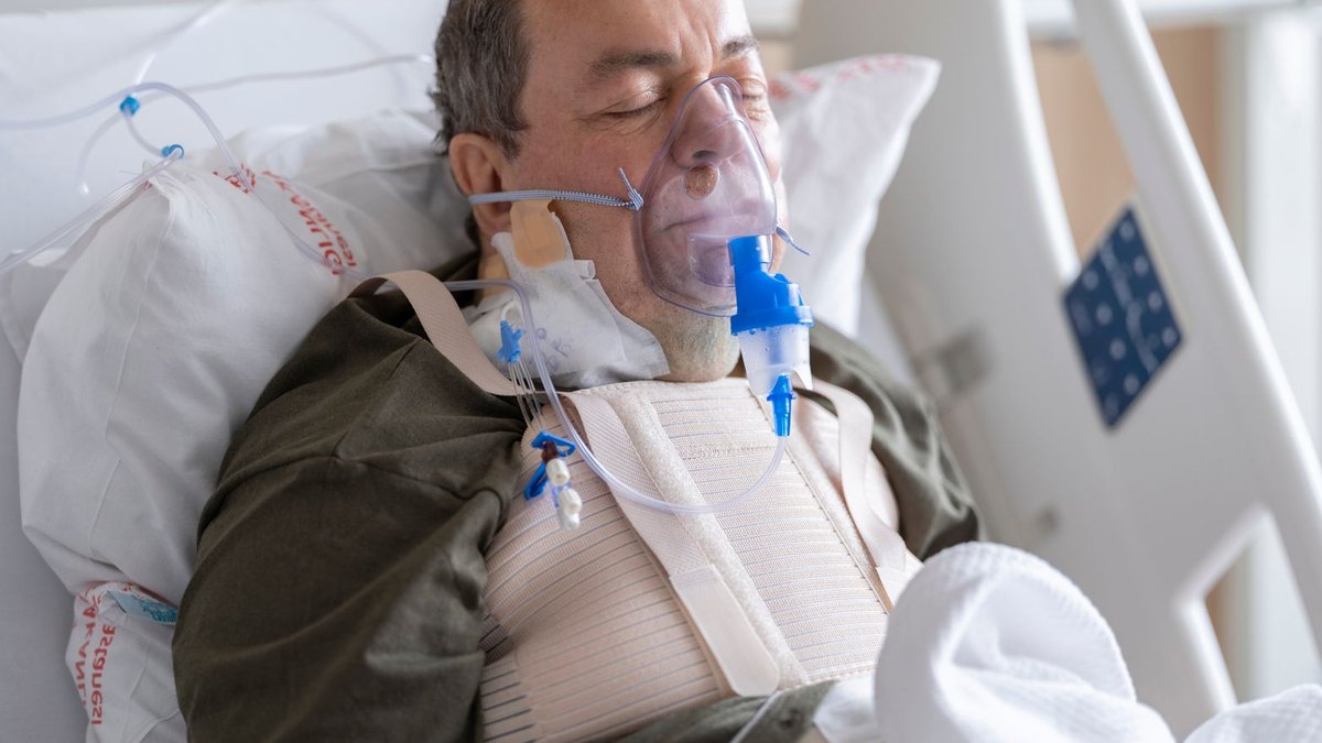 An elderly man lies in a hospital bed wearing an oxygen mask for medical care.