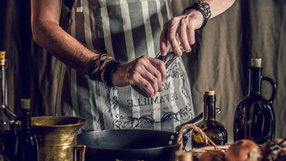 Crop faceless man in apron adding pepper in pan while cooking traditional dish standing at table with assorted ingredient and