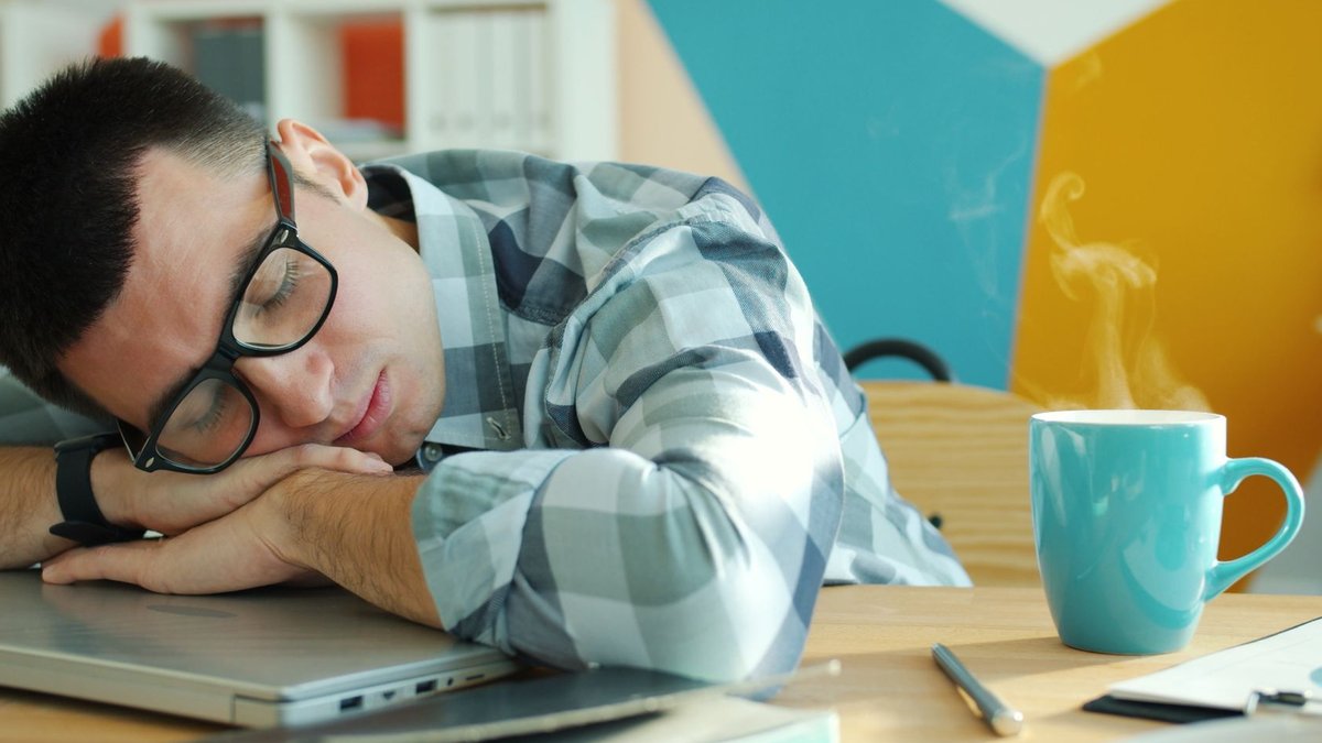 Tired man sleeping on laptop with coffee, showcasing workplace fatigue.