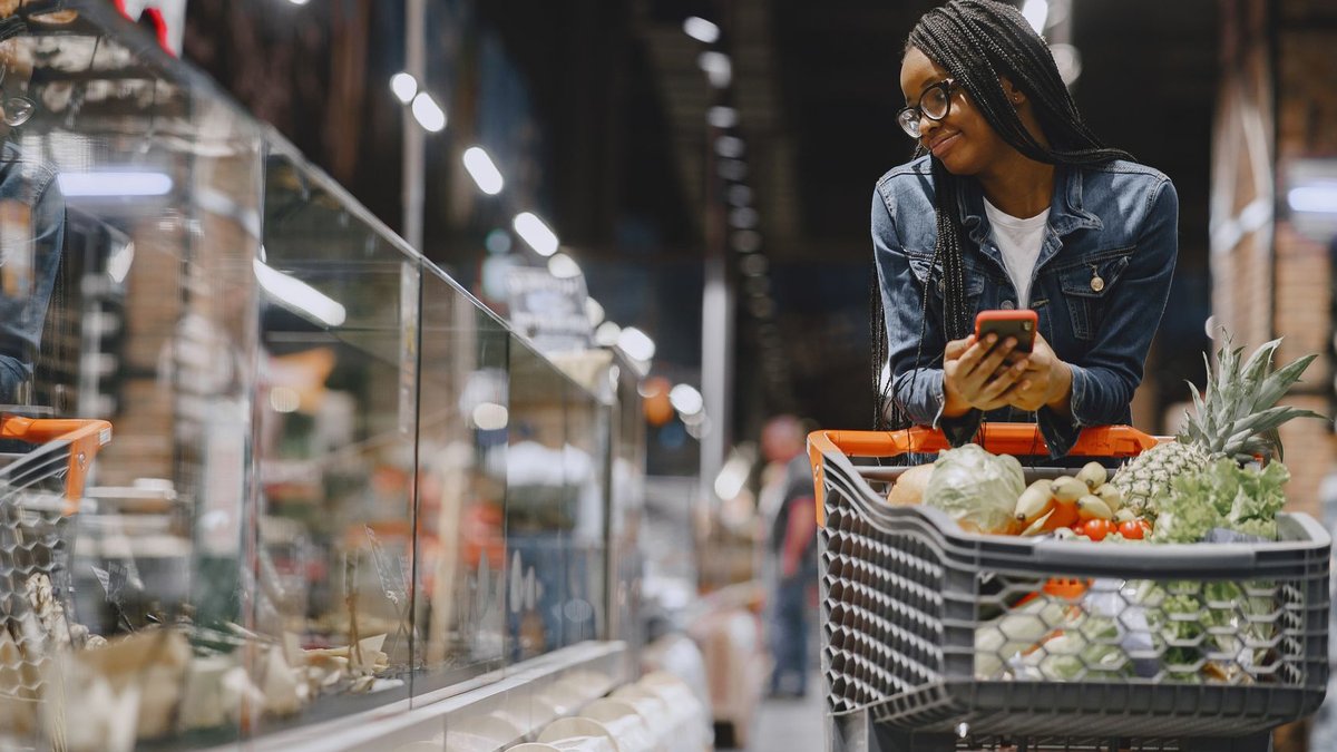 A young woman with afro braids grocery shopping in a supermarket, using a mobile phone.