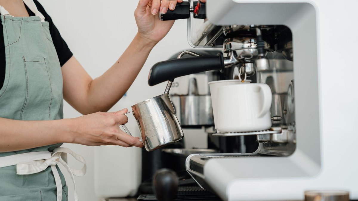 Crop faceless young female barista in apron steaming milk at modern coffee machine while preparing delicious latte.