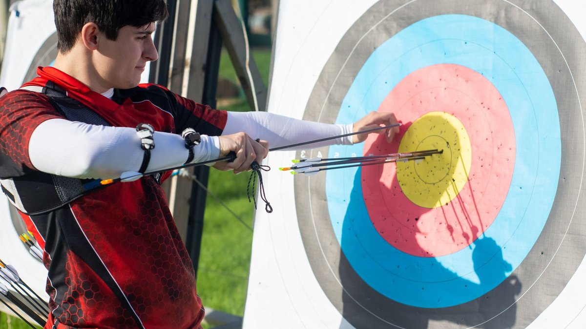 Focus on man aiming with bow and arrows at target during outdoor archery practice.