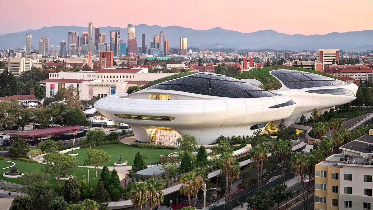 Futuristic white museum buildings with curved roofs sit amid greenery, with a city skyline and mountains in the background at