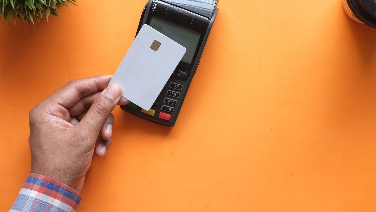 A person making a contactless payment with a credit card and card reader on a bright orange surface.