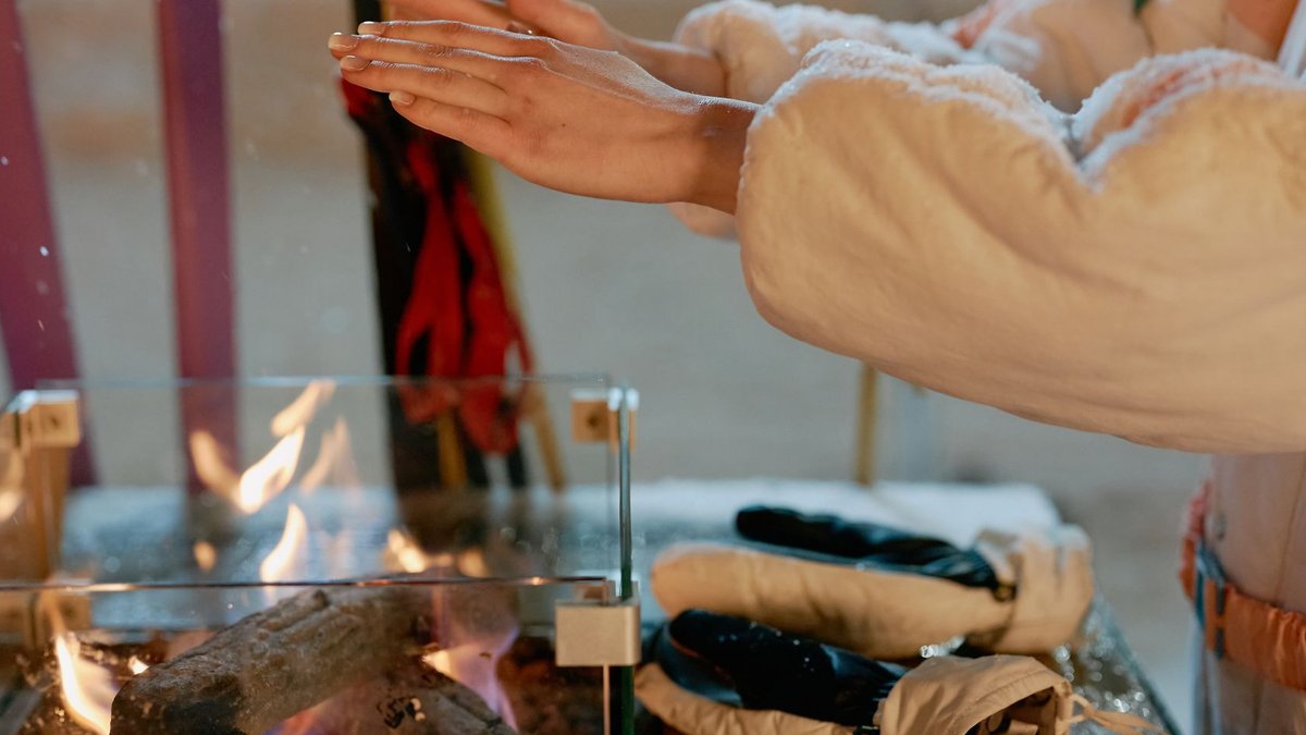 Amazon Offers 45% Off Heated Workwear This Winter Close-up of hands warming by a fire outdoors in winter. Gloves on the table suggest a cold day.