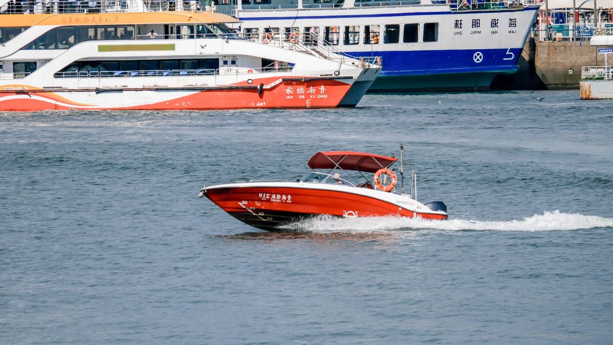 A vibrant speedboat cruising on a river with two ferries docked in the background.
