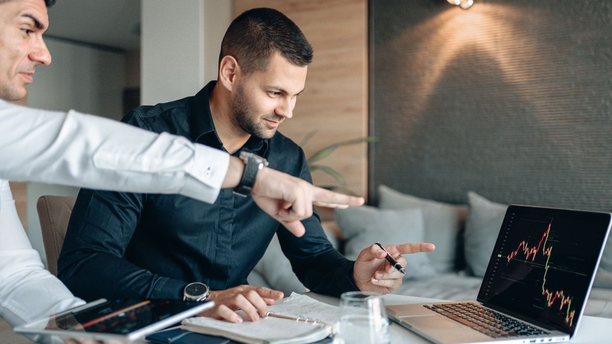 Two businessmen reviewing financial data on a laptop indoors, analyzing market trends.