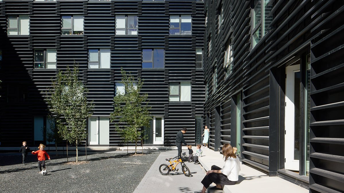 Winnipeg Firm Builds Community Through Innovative Urban Design People and children with a bicycle in a courtyard surrounded by a modern black building with horizontal slats.