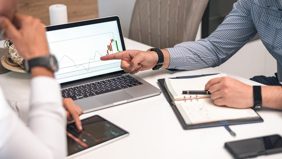 Two businessmen analyzing financial charts and data on a laptop and tablet.