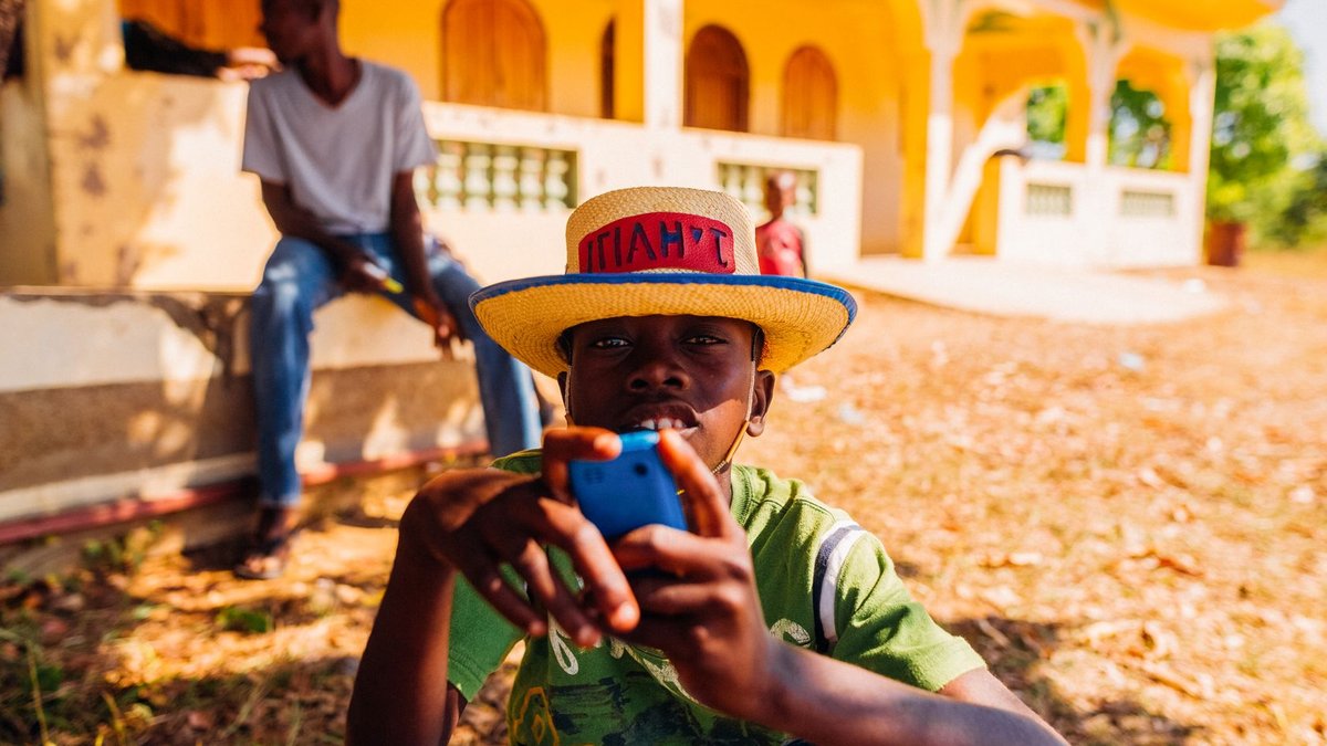 Concentrated African boy in casual clothes and straw hat using mobile phone while resting on yard near house in village on su