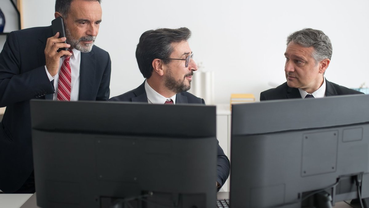Regulator issues AI guidance for pension trustees Three businessmen in an office discussing work, two using computers and one on a phone call.