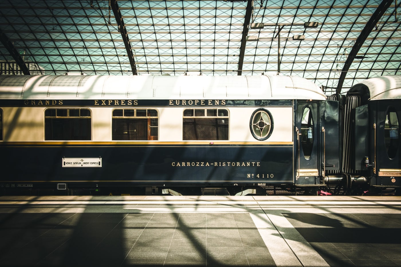 Um vagão-restaurante azul e creme do Venice Simplon-Orient-Express na plataforma, sob a cobertura de vidro de uma estação