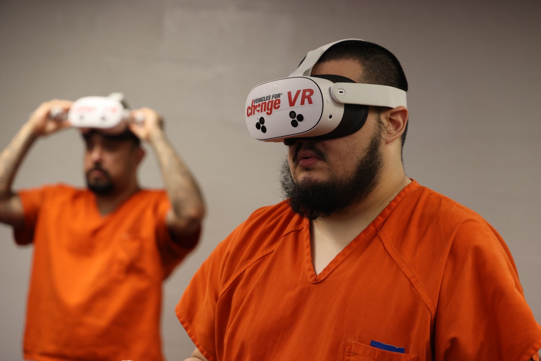A man in an orange jail uniform wears a white VR headset branded Vehicles For Change VR, with a second man behind him lifting a headset onto his head.