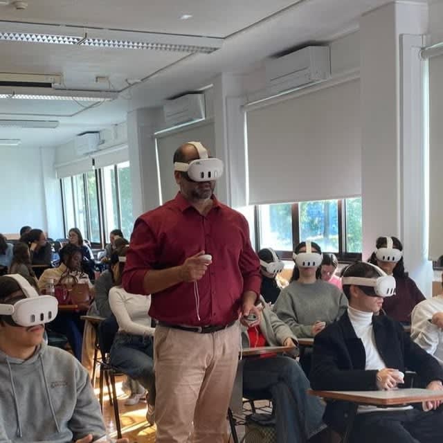 People in a classroom wearing white virtual reality headsets, with a standing participant holding controllers.