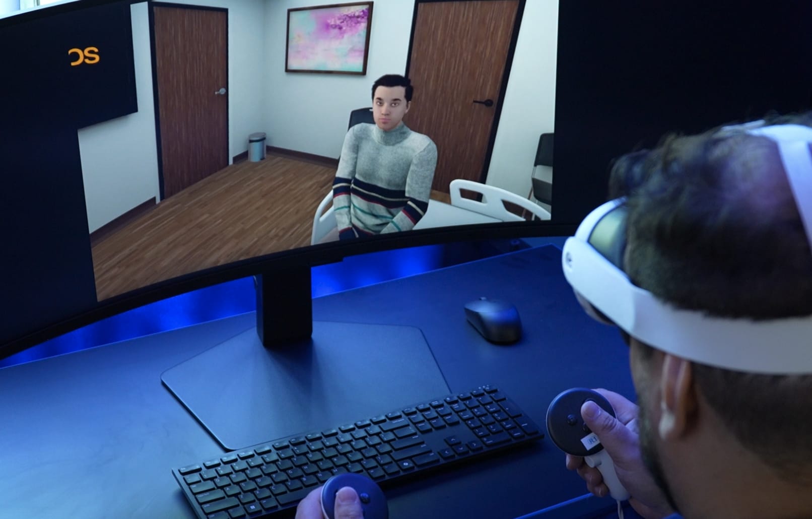 A person wearing a white VR headset holds two controllers at a desk while a curved monitor shows a virtual client seated in a simulated counseling office.