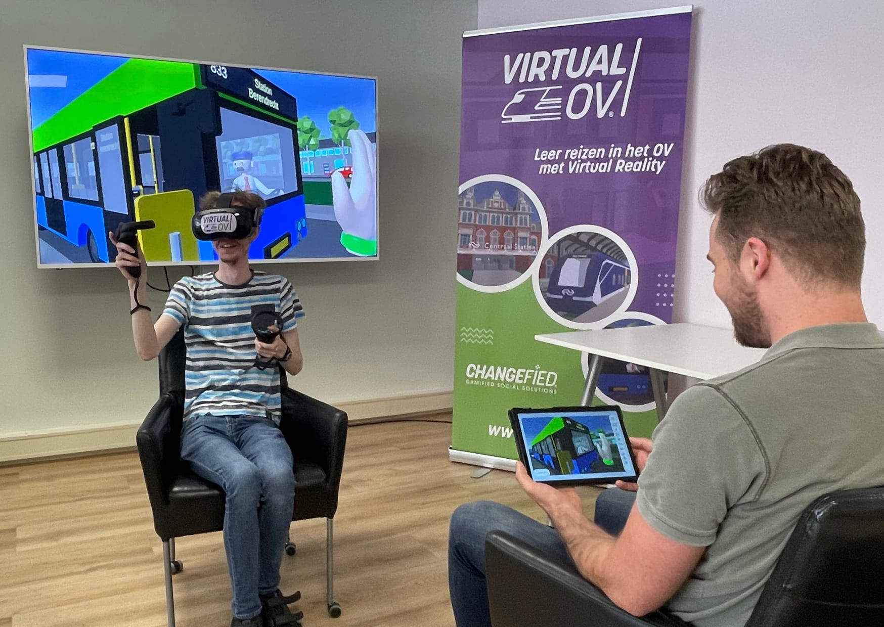 A person in a Virtual OV VR headset with controllers practises a bus journey, the scene mirrored on a screen behind, while a facilitator follows on a tablet.
