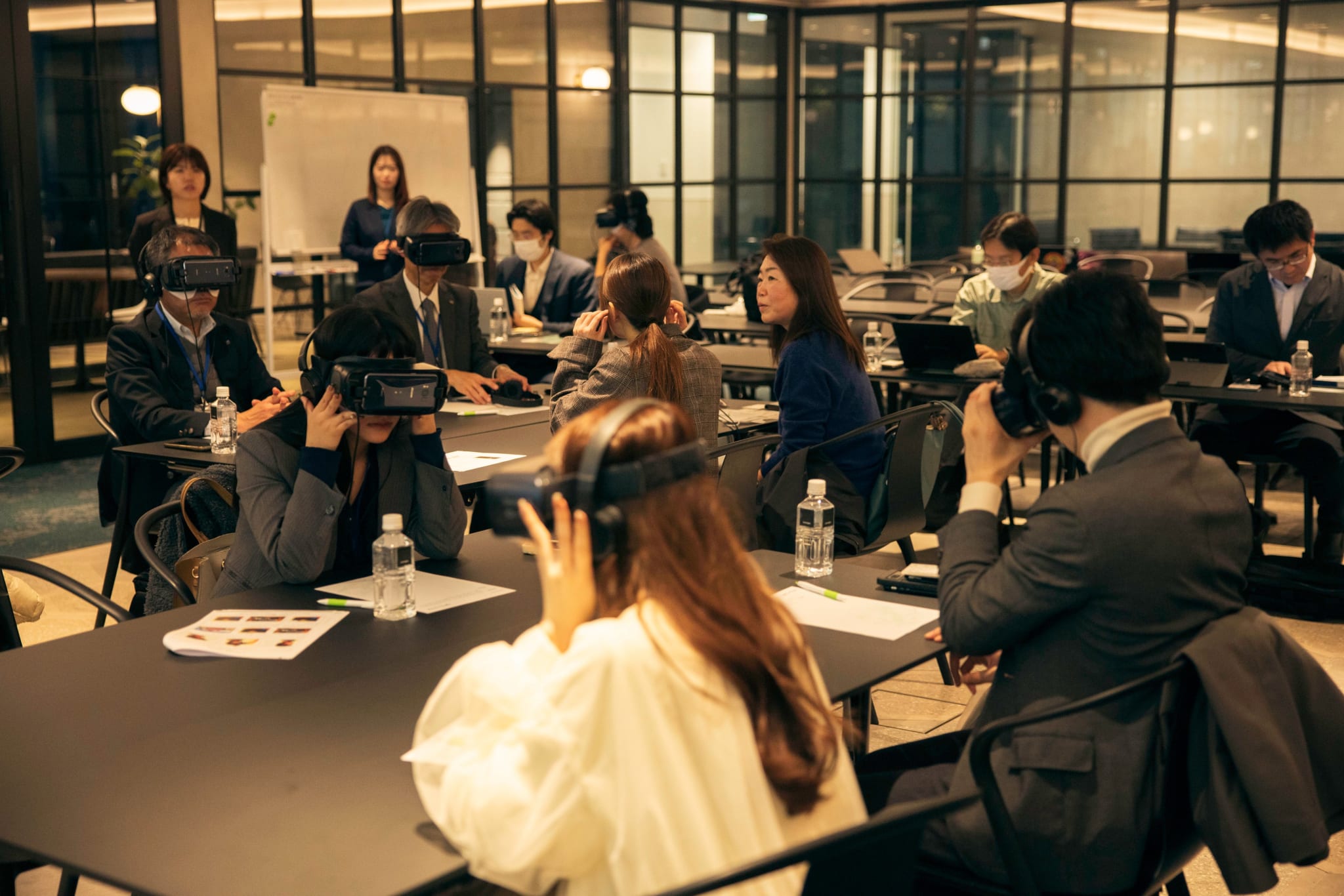 About a dozen seminar participants seated at meeting-room tables, each wearing a VR viewer and over-ear headphones during the first-person simulation.