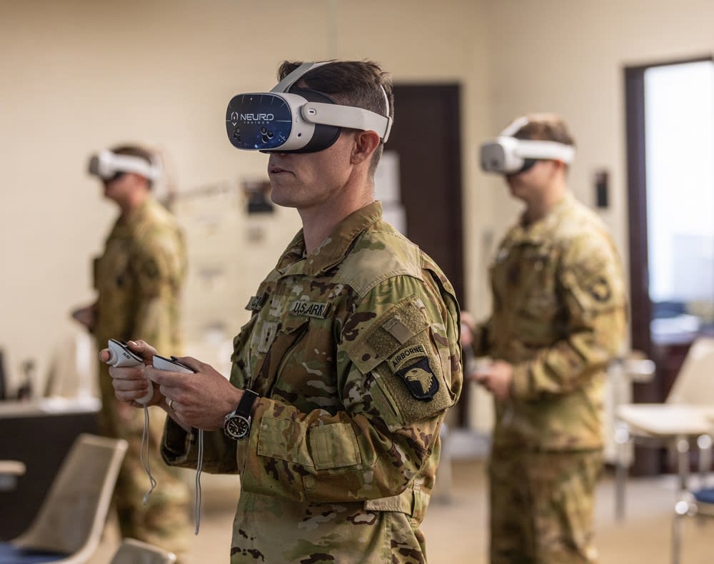 Three U.S. Army Soldiers in camouflage uniforms stand in a classroom wearing white VR headsets branded NeuroTrainer, holding motion controllers; the foreground Soldier wears a 101st Airborne patch.