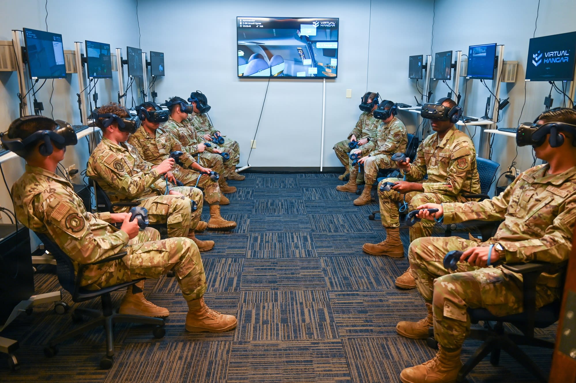Two rows of Airmen in camouflage uniforms face each other across a carpeted VR lab, each seated in a VR headset holding motion controllers, with monitors along both walls, a centre screen showing a C-5M Aircraft Egress scene and a Virtual Hangar logo display on the right.