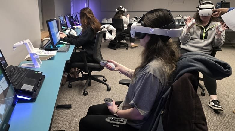 A Carroll Community College student uses a VR headset and controllers in the college's VR lab while classmates use headsets in the background.
