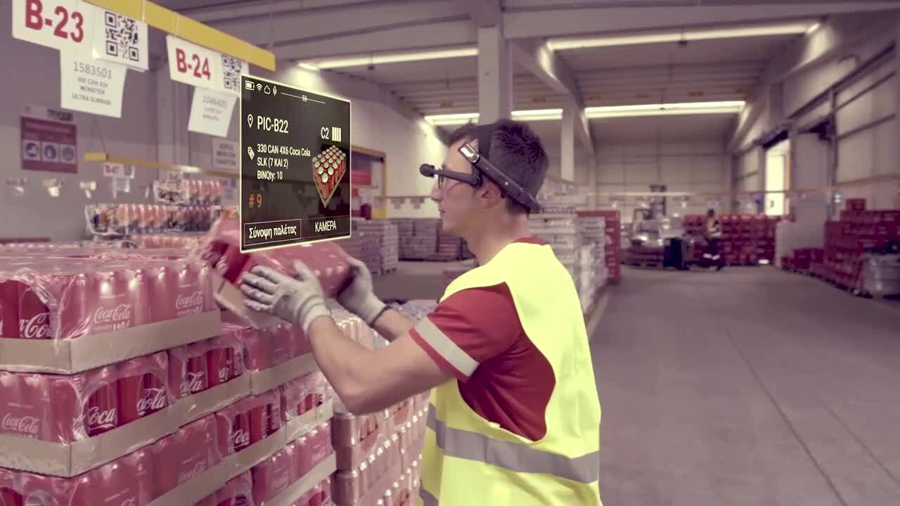 A warehouse picker in a hi-vis vest wearing a head-mounted display lifts a tray of Coca-Cola cans onto a pallet, with the Greek-language picking screen showing the aisle code, product and quantity.
