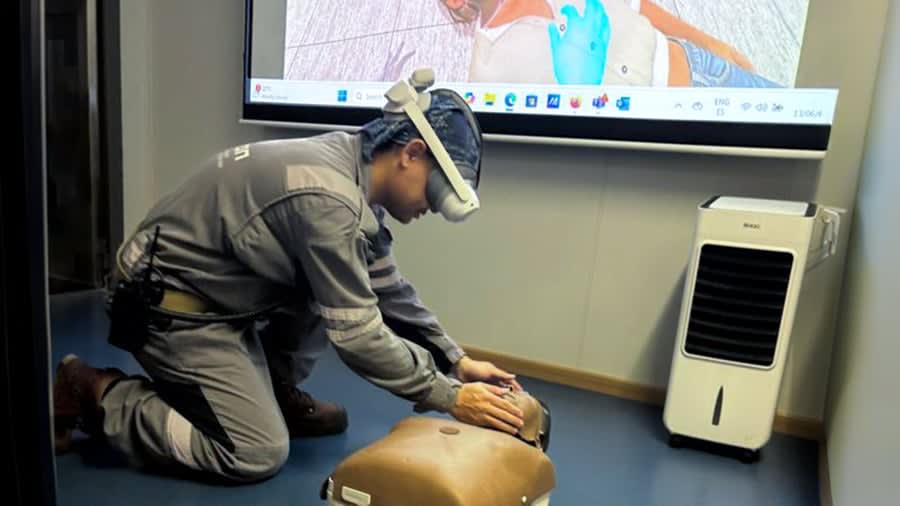 Drillship crew member in grey coveralls kneels beside a CPR manikin while wearing a standalone VR headset, the simulation mirrored on a screen behind him.