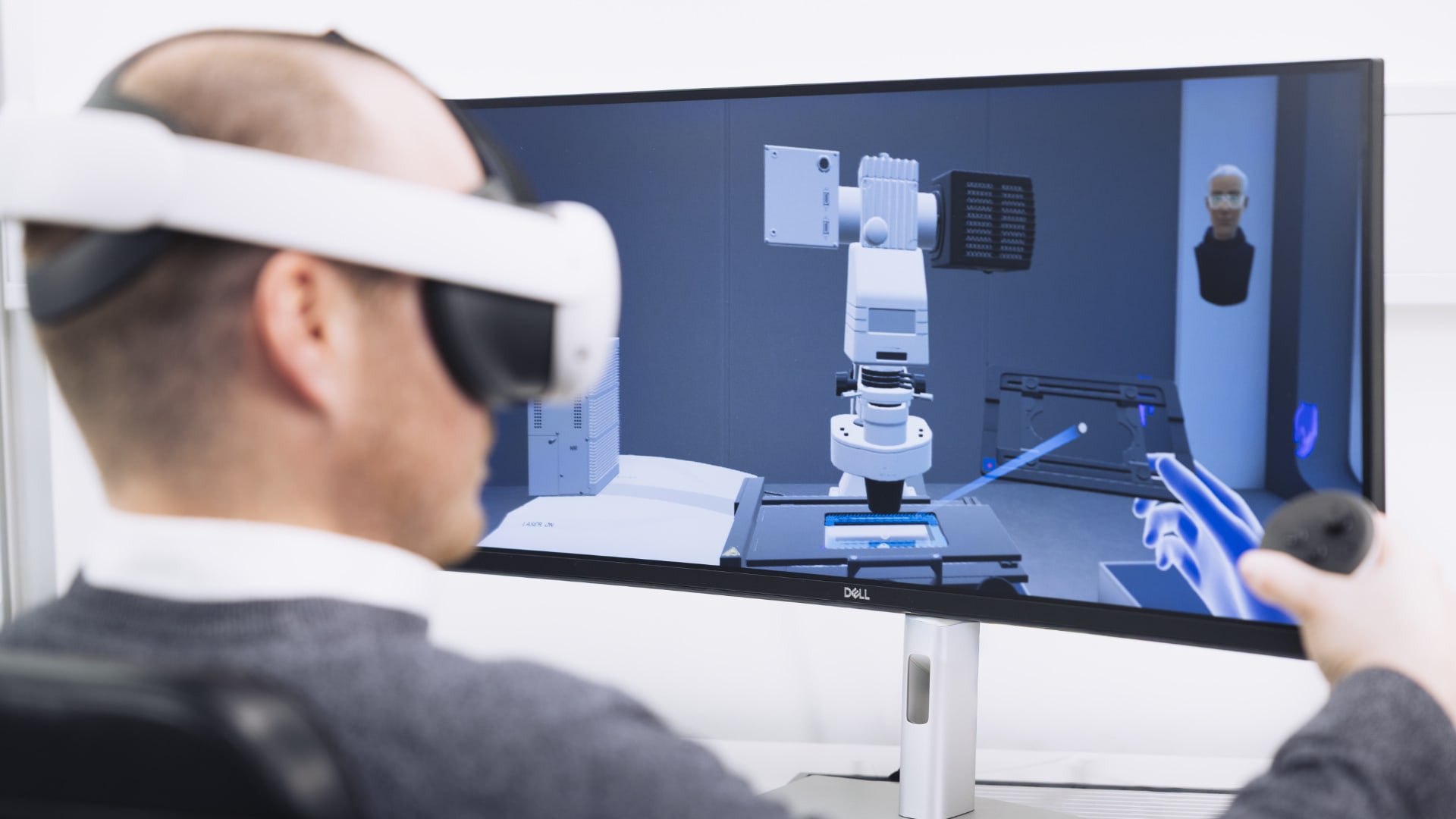 A person is wearing a virtual reality headset and is sitting at a desk, holding a controller in their right hand. The display on the desk shows the virtual environment the person is participating in.