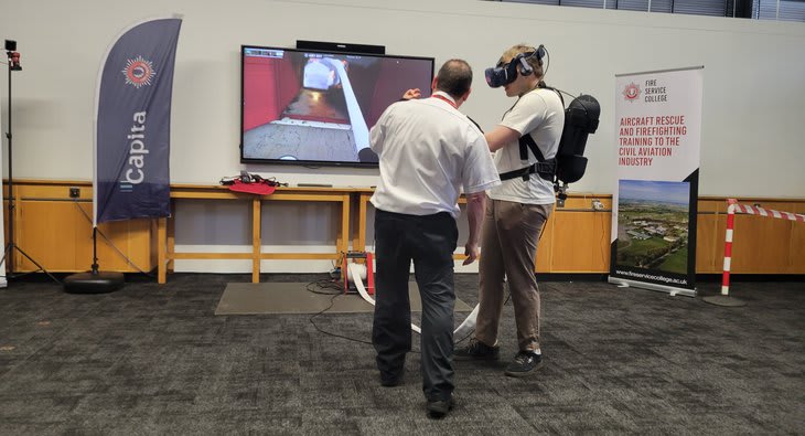 A trainee wearing a VR headset and a backpack harness holds a hose controller beside an instructor, with the virtual fire scene shown on a wall screen and Fire Service College banners behind them.