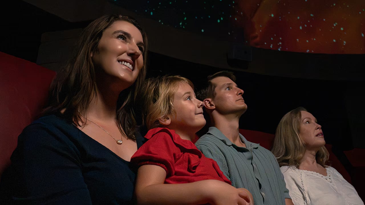 A woman, a young girl, a man and an older woman sit in red seats looking up at stars projected on the dome overhead.