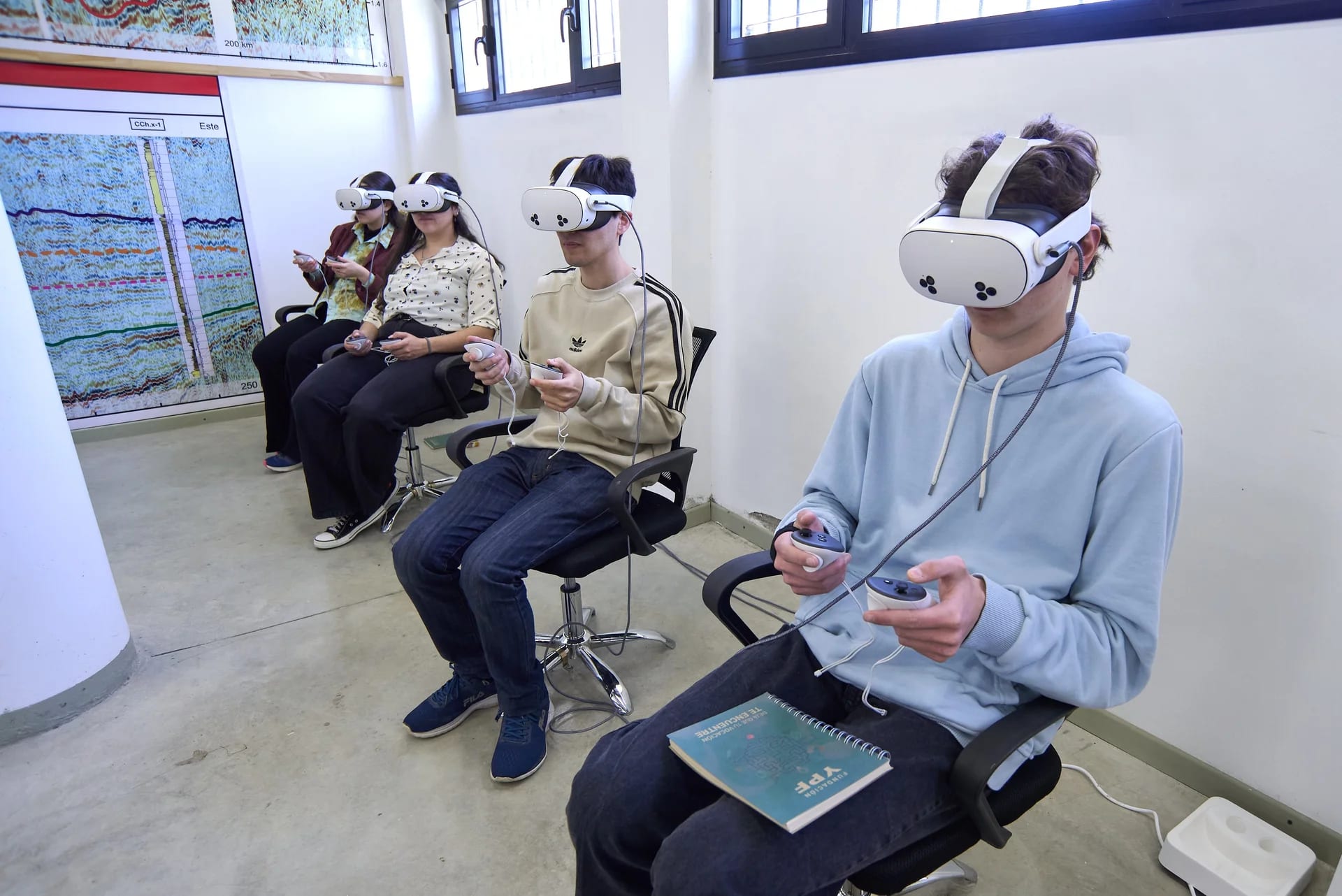 Four secondary students seated in a row wearing white VR headsets and holding controllers, with a seismic-section poster on the wall beside them.