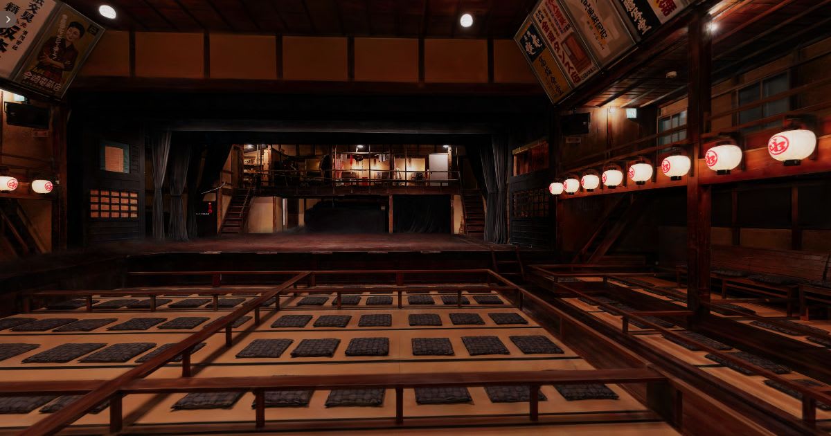 The Eirakukan auditorium seen from the rear of the tatami box seating, looking across to the lit stage beneath rows of paper lanterns and painted advertising boards