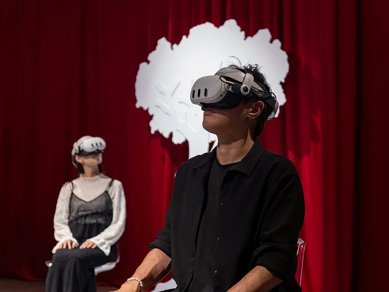 Two visitors seated before a red curtain wearing white VR headsets