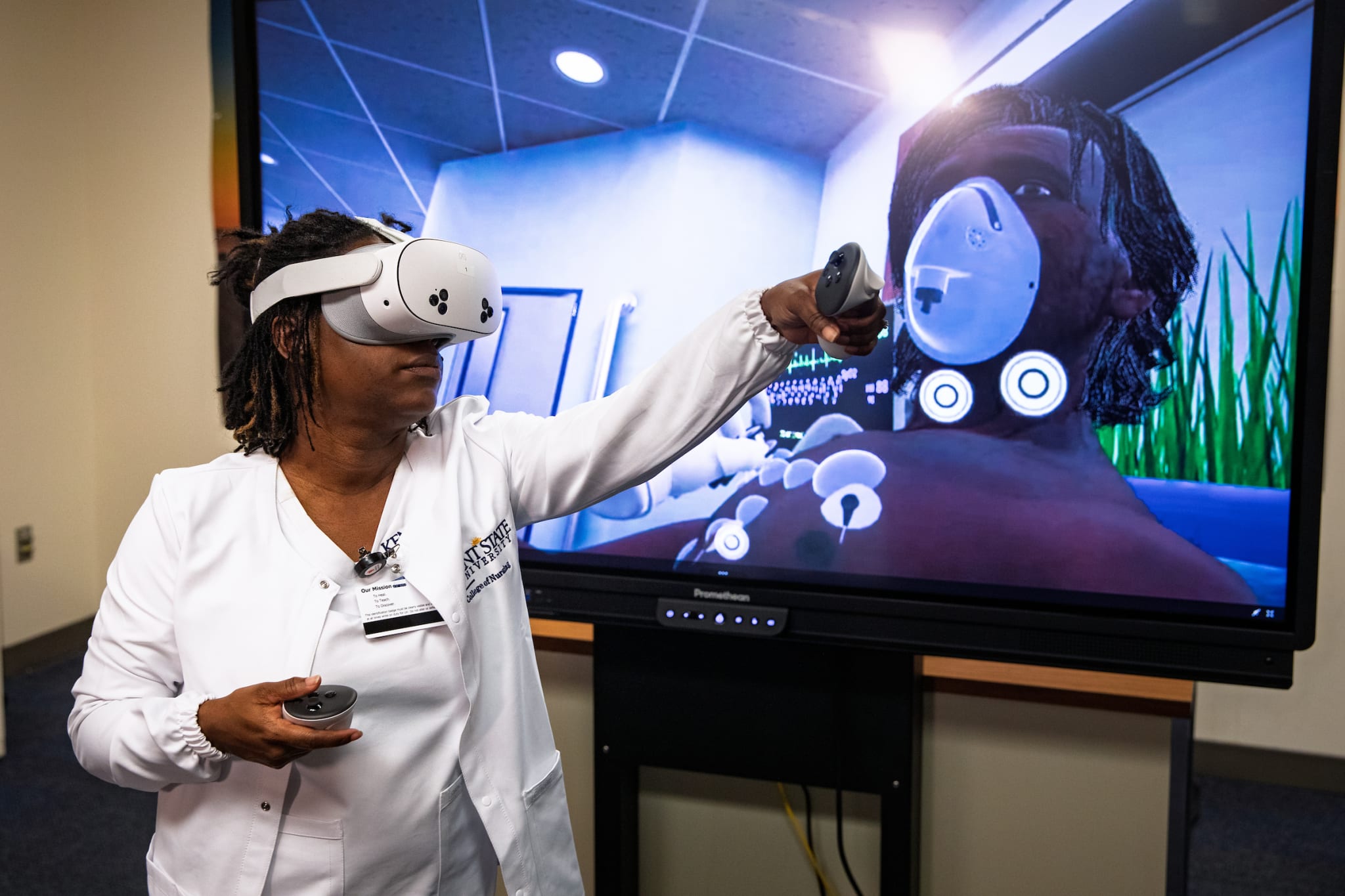 A Kent State nursing student wearing a VR headset holds controllers beside a screen displaying a virtual patient wearing an oxygen mask.