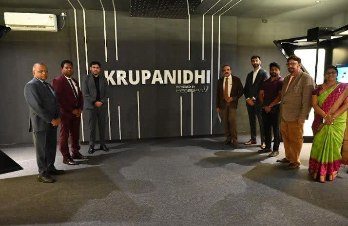 Nine people in formal dress stand in the new lab in front of an illuminated wall sign reading KRUPANIDHI, powered by MediSim VR, with a white VR training station at the right of frame.