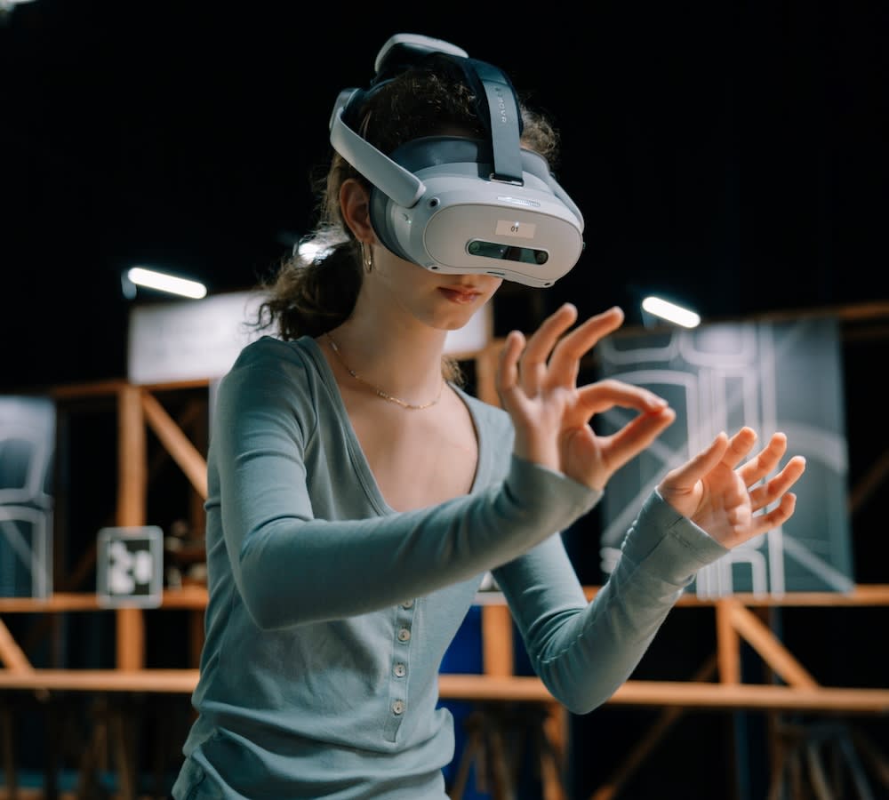 A visitor wearing a white standalone headset gestures with both hands in the dimly lit XR Workshop room of the Gaudí Code exhibition, with wooden workbench frames and Gaudí drawings behind her.