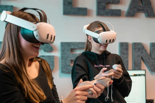 Two teenage girls wearing white standalone VR headsets and holding hand controllers, smiling as they use a virtual reality experience in front of a wall with large cut-out lettering.