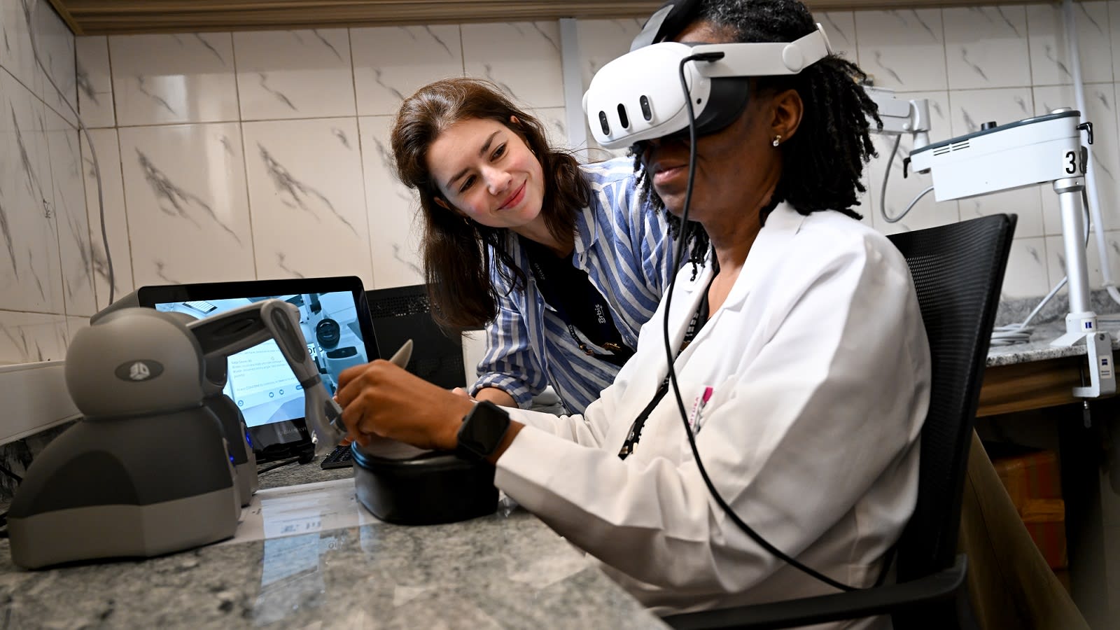 An ophthalmology trainee in a white coat wears a VR headset and guides the stylus of a desktop haptic arm while an Orbis colleague watches the cataract simulation on the laptop beside them.