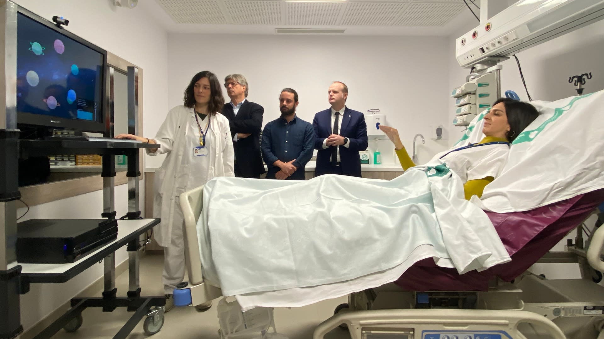 A woman raises her hand from a hospital bed toward a screen showing colorful planets while a clinician and three observers stand beside the equipment.