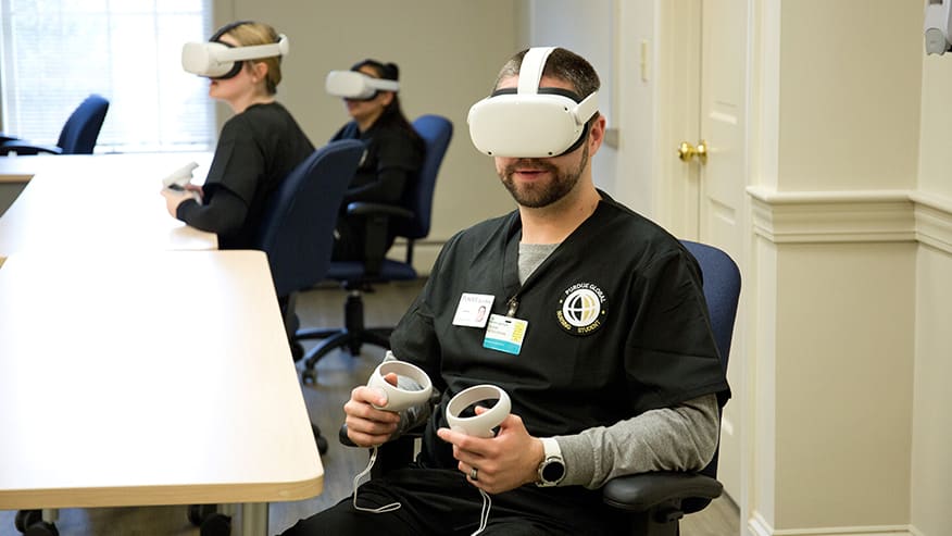 Three Purdue Global nursing students sit at a table wearing VR headsets and holding handheld controllers